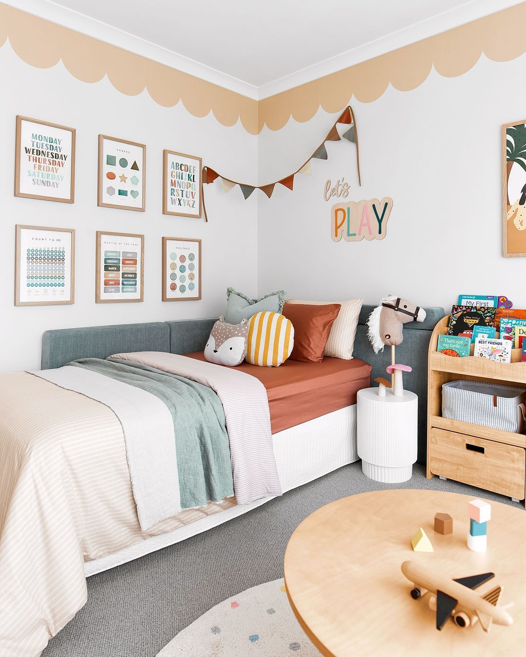 A cozy children's bedroom with a bed featuring multi-colored bedding and decorative pillows. The walls are adorned with framed educational posters and a playful scalloped wallpaper border in a soft peach color near the ceiling.