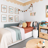 A cozy children's bedroom with a bed featuring multi-colored bedding and decorative pillows. The walls are adorned with framed educational posters and a playful scalloped wallpaper border in a soft peach color near the ceiling.