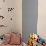 Children's reading nook with a cozy floor pillow and a blue sticker arch on the wall. The wall features playful farm animal decals, and a wooden bookshelf displays various children's books