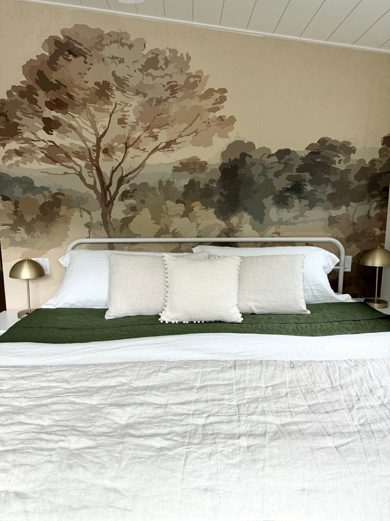A serene bedroom featuring a Vintage Landscape mural with a tree dominating the scene, flanked by gold lamps and crisp white bedding with a green accent.