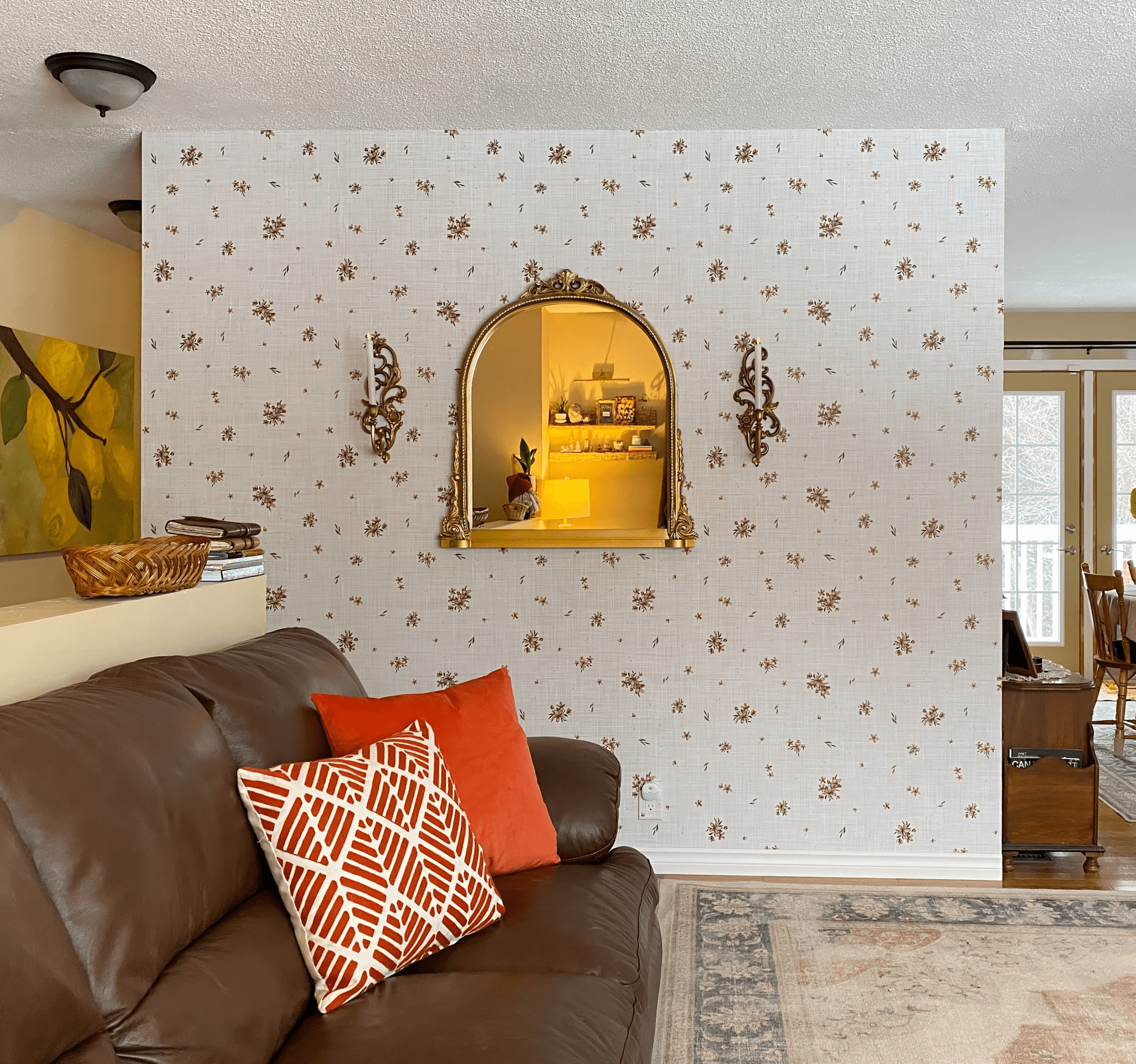 A room with a wall covered in beige wallpaper featuring a floral motif with brown and pink flowers. A gold-framed mirror and ornate sconces are on the wall.