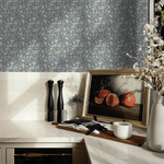A stylish kitchen corner with blue vintage floral wallpaper. The countertop displays elegant salt and pepper mills, a framed picture of pomegranates, a ceramic pitcher with blossoming branches, and a modern candle holder