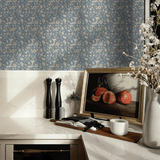 A stylish kitchen corner with blue vintage floral wallpaper. The countertop displays elegant salt and pepper mills, a framed picture of pomegranates, a ceramic pitcher with blossoming branches, and a modern candle holder