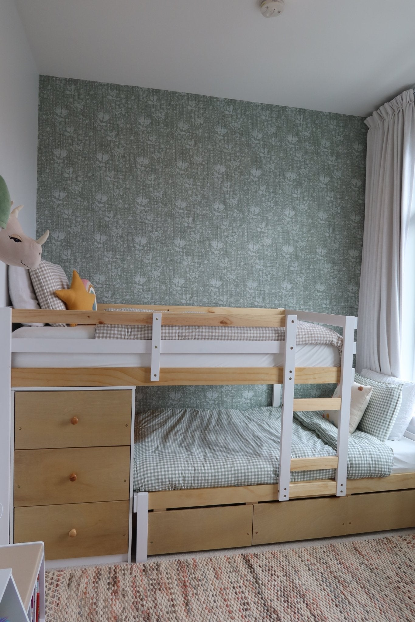 Bright and airy child's bedroom with pine bunk bed, forest green floral wallpaper, and a window draped with sheer white curtains.