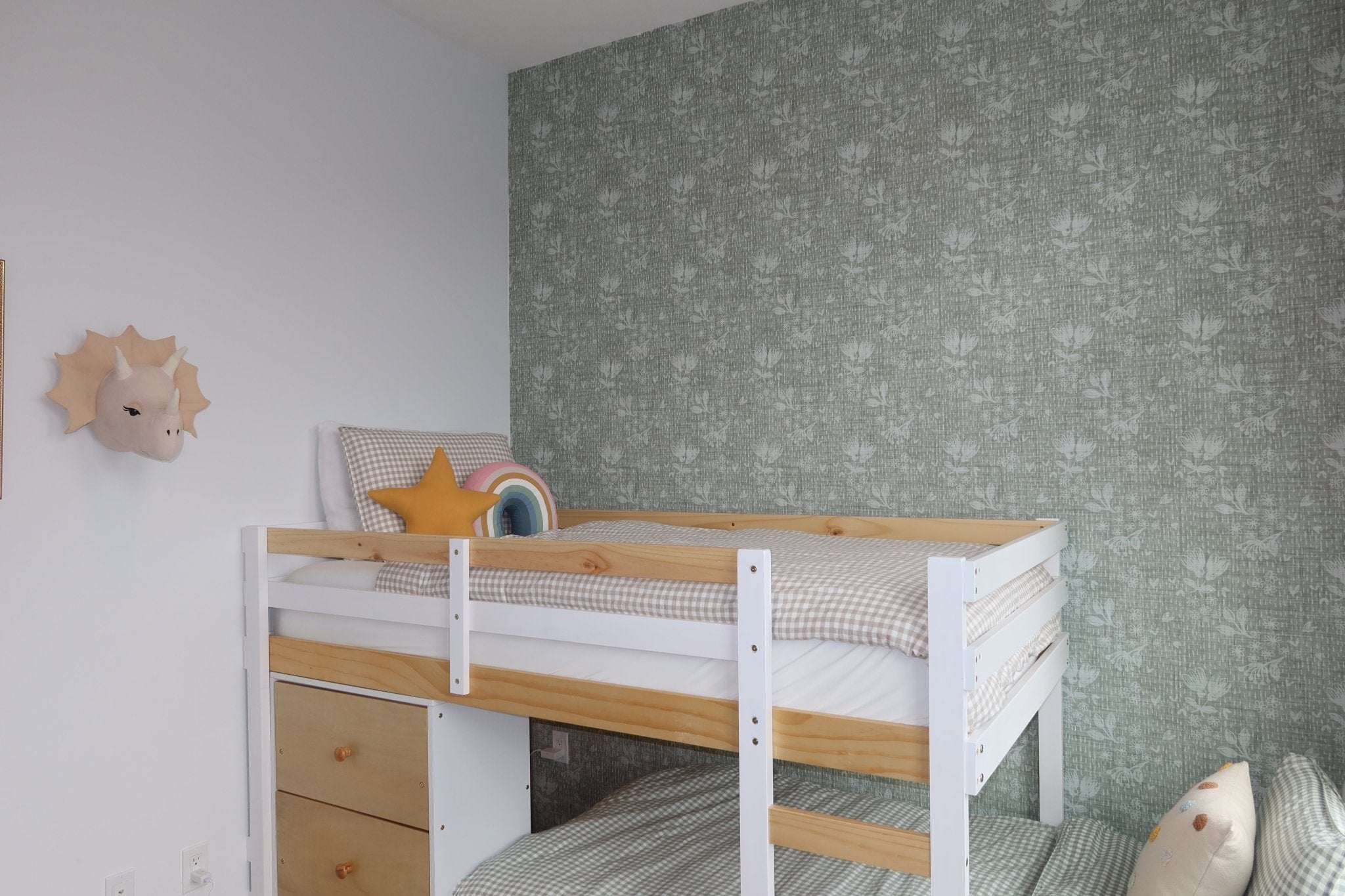 A child's bedroom featuring a white and pine bunk bed against a forest green wallpaper with a subtle floral pattern and a stuffed triceratops head wall decor.
