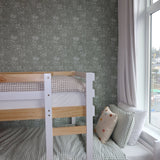 Pine bunk bed with white accents in a room with forest green floral wallpaper, adjacent to a window with sheer white curtains.