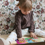 A young child sits on a cushioned floor seat, reading a book against a backdrop of floral wallpaper with maroon and olive heirloom-style flowers
