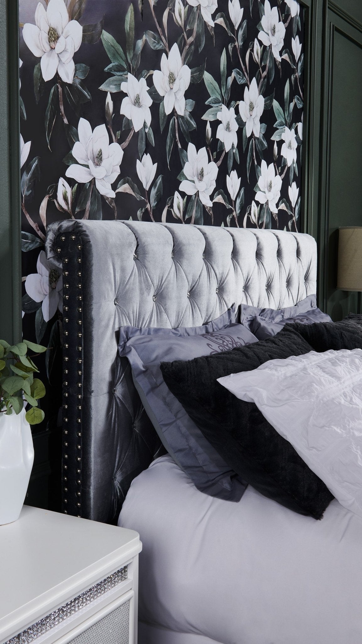 A close-up of a bedroom showing a wallpaper with white magnolias on a black background, matching the gray tufted headboard with black accents.