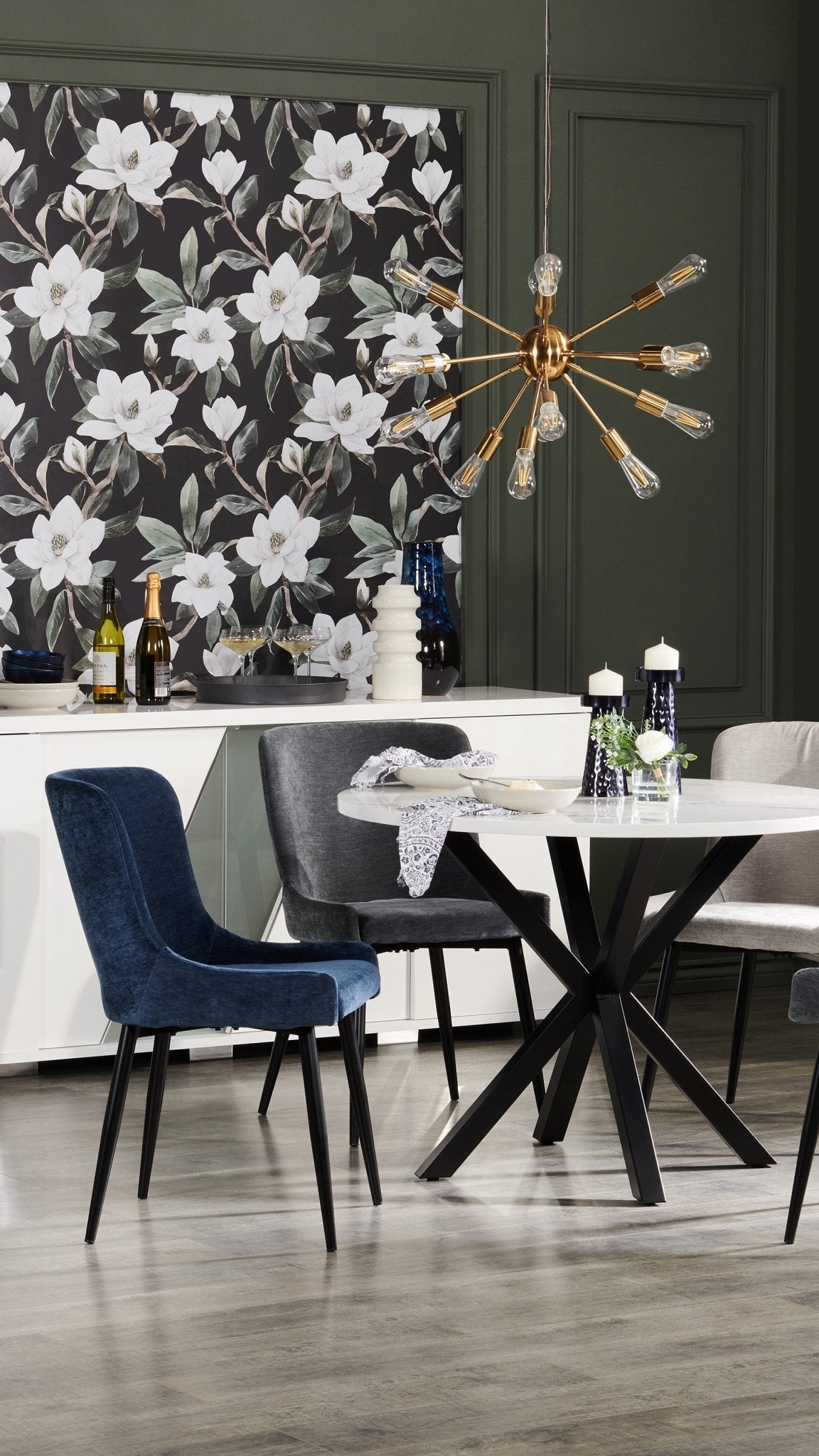 A modern dining area with the same magnolia wallpaper, which acts as a bold backdrop to a white table, blue velvet chairs, and a striking gold light fixture.