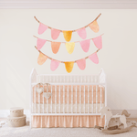 A nursery room with a white crib adorned with two heart-shaped ornaments. Above the crib, peel-and-stick bunting banners in pastel pink and yellow hues are displayed on the wall. To the crib's left are two stacked round storage boxes, and to the right is a wooden rocking horse on a rug