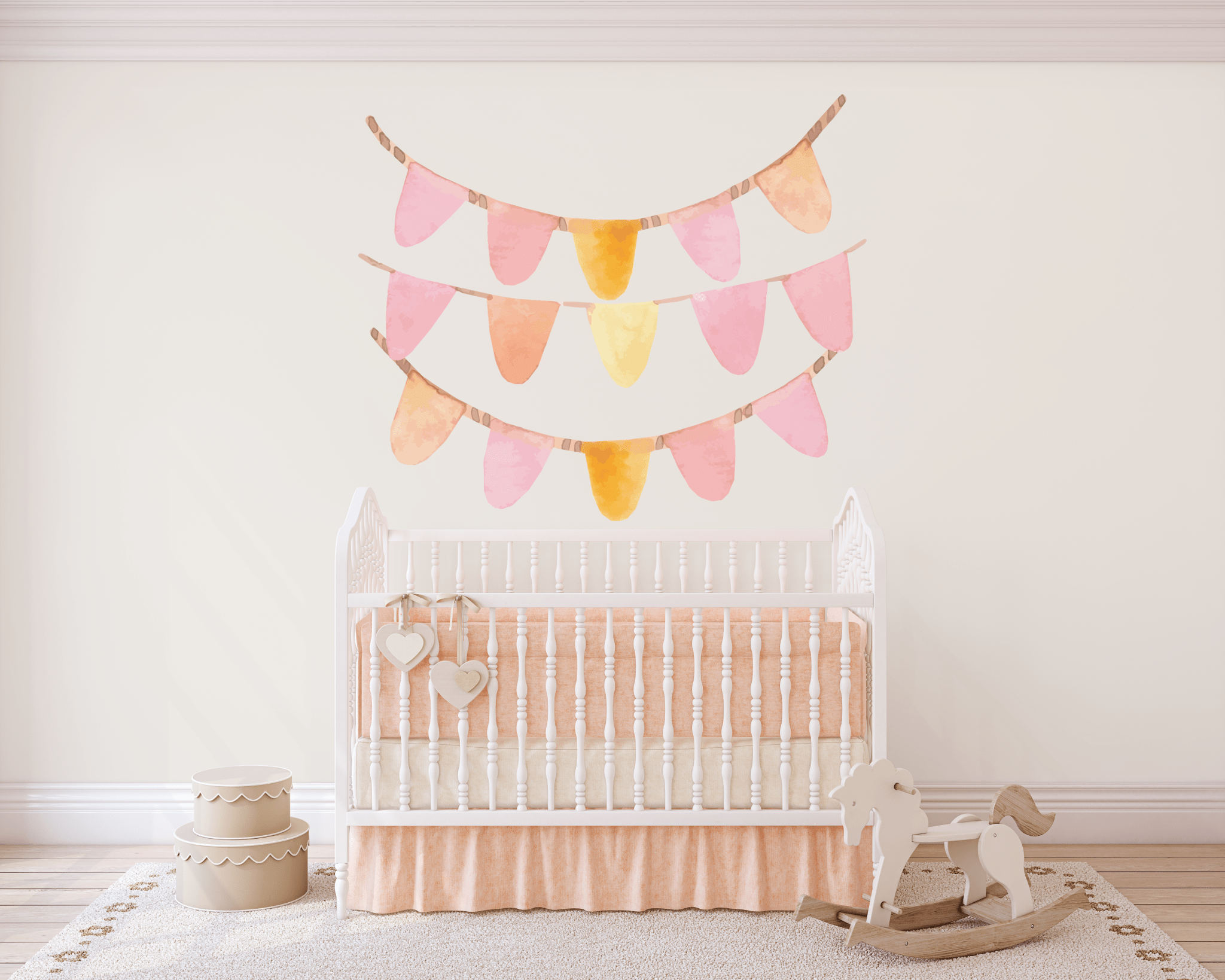 A nursery room with a white crib adorned with two heart-shaped ornaments. Above the crib, peel-and-stick bunting banners in pastel pink and yellow hues are displayed on the wall. To the crib's left are two stacked round storage boxes, and to the right is a wooden rocking horse on a rug