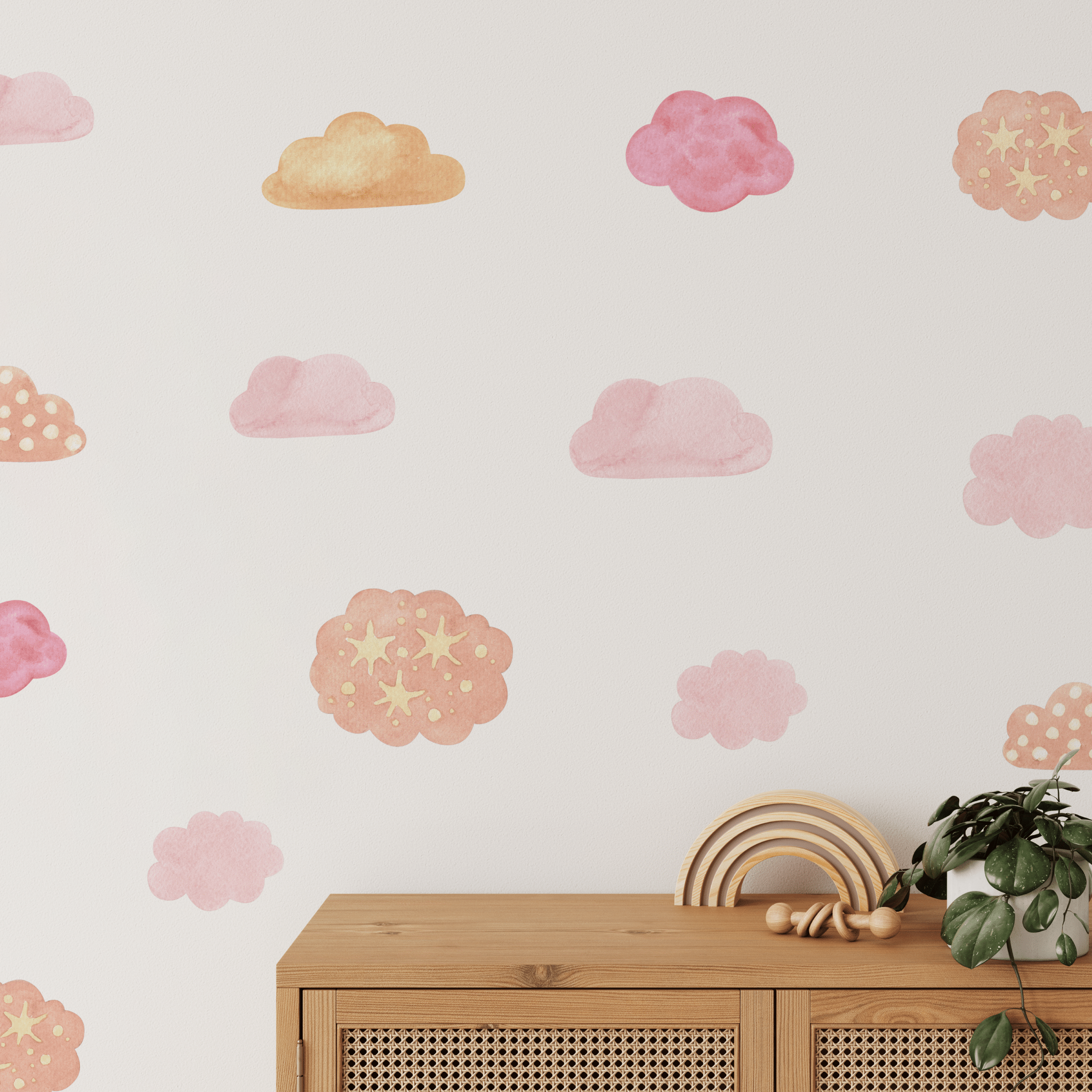 A nursery room wall decorated with watercolor wall stickers of clouds in shades of pink and gold, with a wooden toy rainbow on a cabinet and a hanging plant