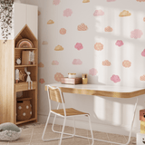 A nursery room wall decorated with watercolor wall stickers of clouds in shades of pink and gold, with a wooden toy rainbow on a cabinet and a hanging plant."