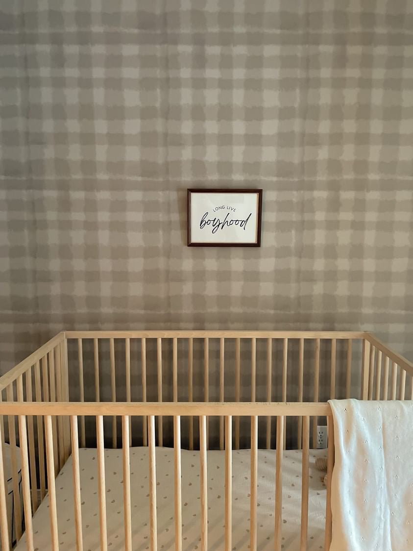 Minimalist nursery with a wooden crib, stuffed toy, and wall art reading "long live boyhood" against plaid wallpaper.