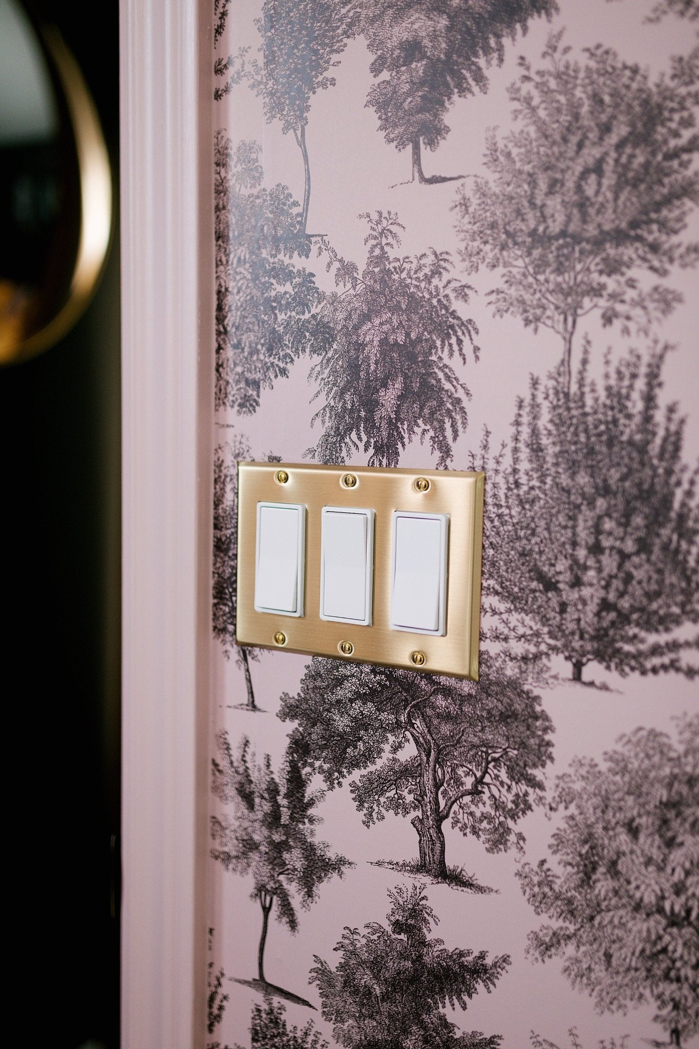 A light switch with three white switches on a brass plate against a toile wallpaper with pink trees and foliage.