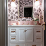 A bathroom vanity with white cabinets, brass handles, and a pink toile tree-pattern wallpaper.