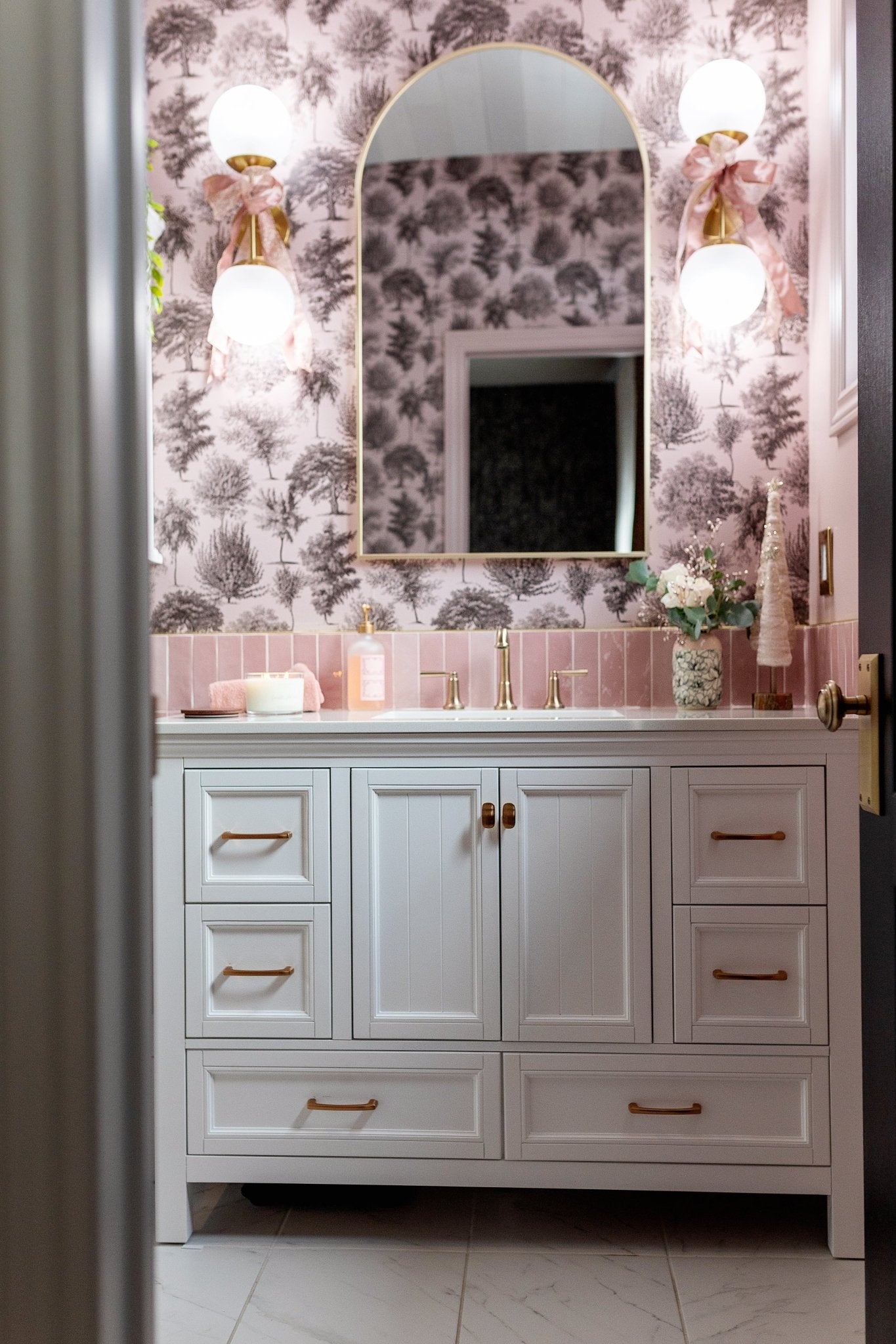 A bathroom vanity with white cabinets, brass handles, and a pink toile tree-pattern wallpaper.