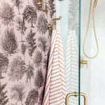 A shower with pink toile tree-pattern wallpaper, brass hooks holding striped towels, and a glass door.