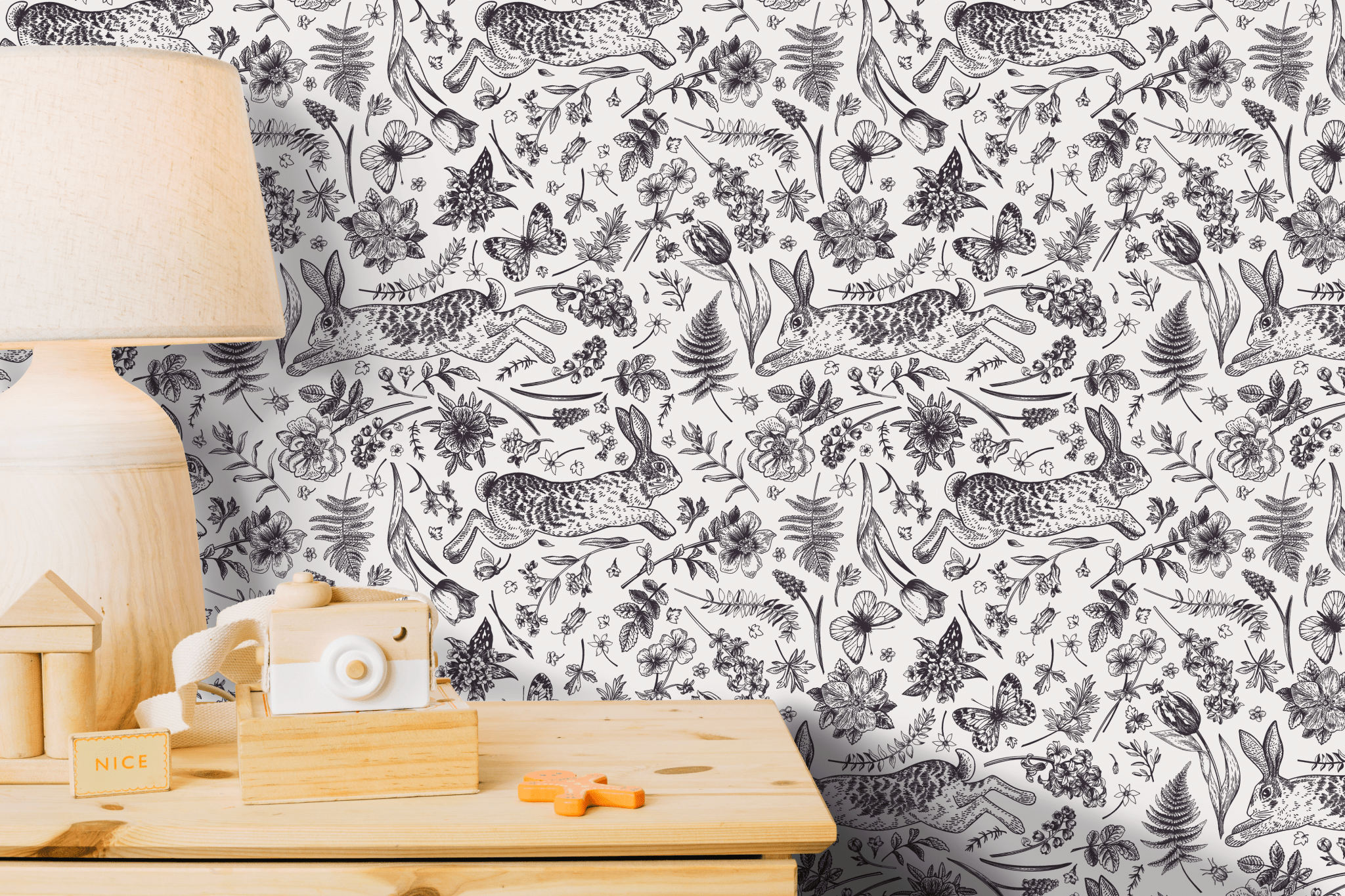 A close-up view of peel and stick wallpaper featuring an intricate hand-drawn style pattern of rabbits, tulips, butterflies, and ferns in a monochromatic black and white color scheme on a wall behind a wooden table with a lamp and toy camera