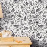 A close-up view of peel and stick wallpaper featuring an intricate hand-drawn style pattern of rabbits, tulips, butterflies, and ferns in a monochromatic black and white color scheme on a wall behind a wooden table with a lamp and toy camera