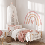 A cozy corner of a child's bedroom featuring a pastel rainbow wall decal. The room includes a white canopy over a single bed with pink bedding, a fluffy white rug on the floor, and a small wooden desk with a red chair.