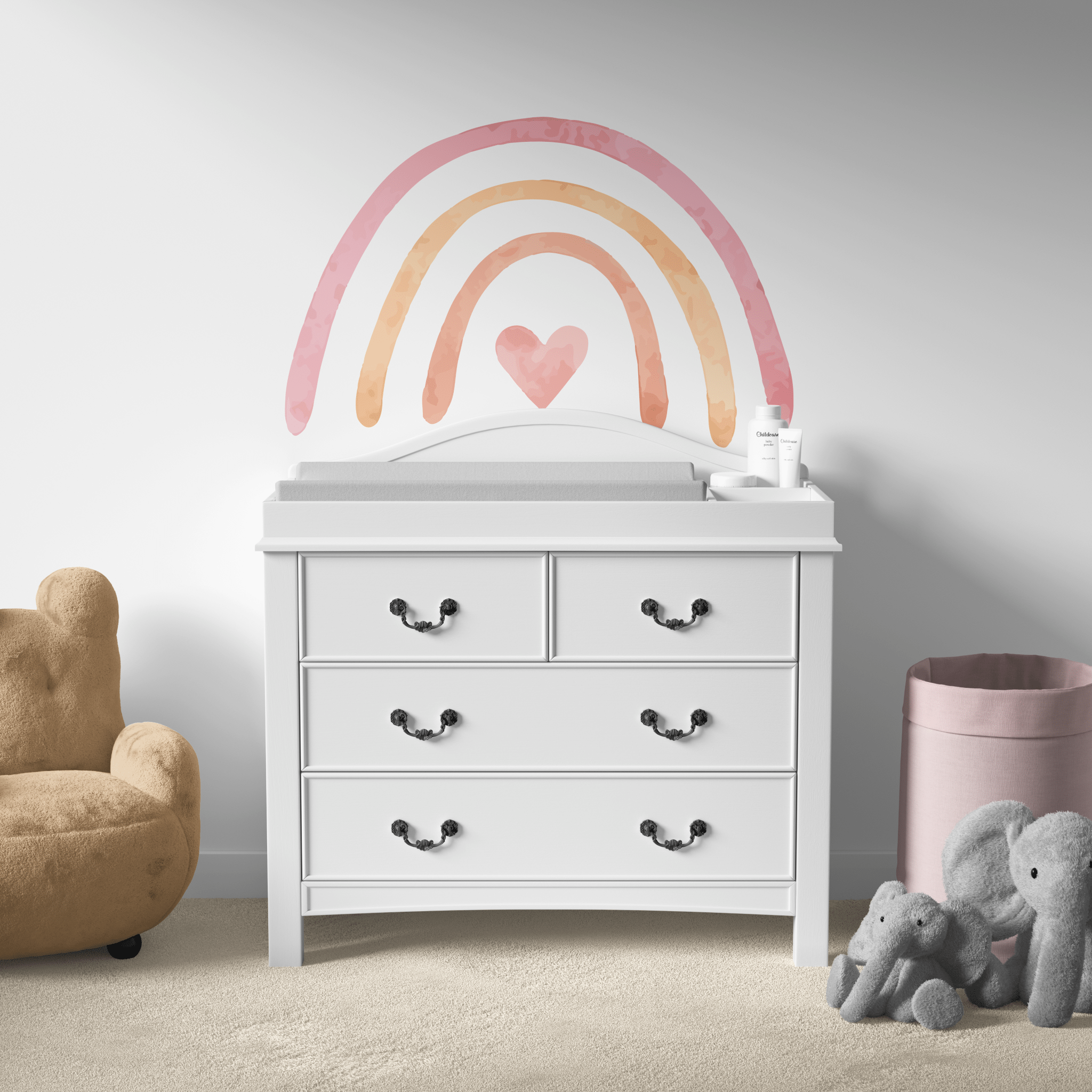 A simple and modern nursery setup with a pastel rainbow wall decal above a white changing table. The room includes a comfy brown chair, a soft elephant toy, and a pink storage bin, creating a gentle and relaxing atmosphere.