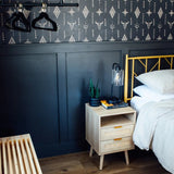  Another angle of the bedroom highlighting the Western-inspired wallpaper complemented by a dark blue paneled wall below. Beside the bed with crisp white linens is a natural wood bedside table with rattan drawer fronts, topped with a modern lantern-style lamp, books, and a small potted plant.