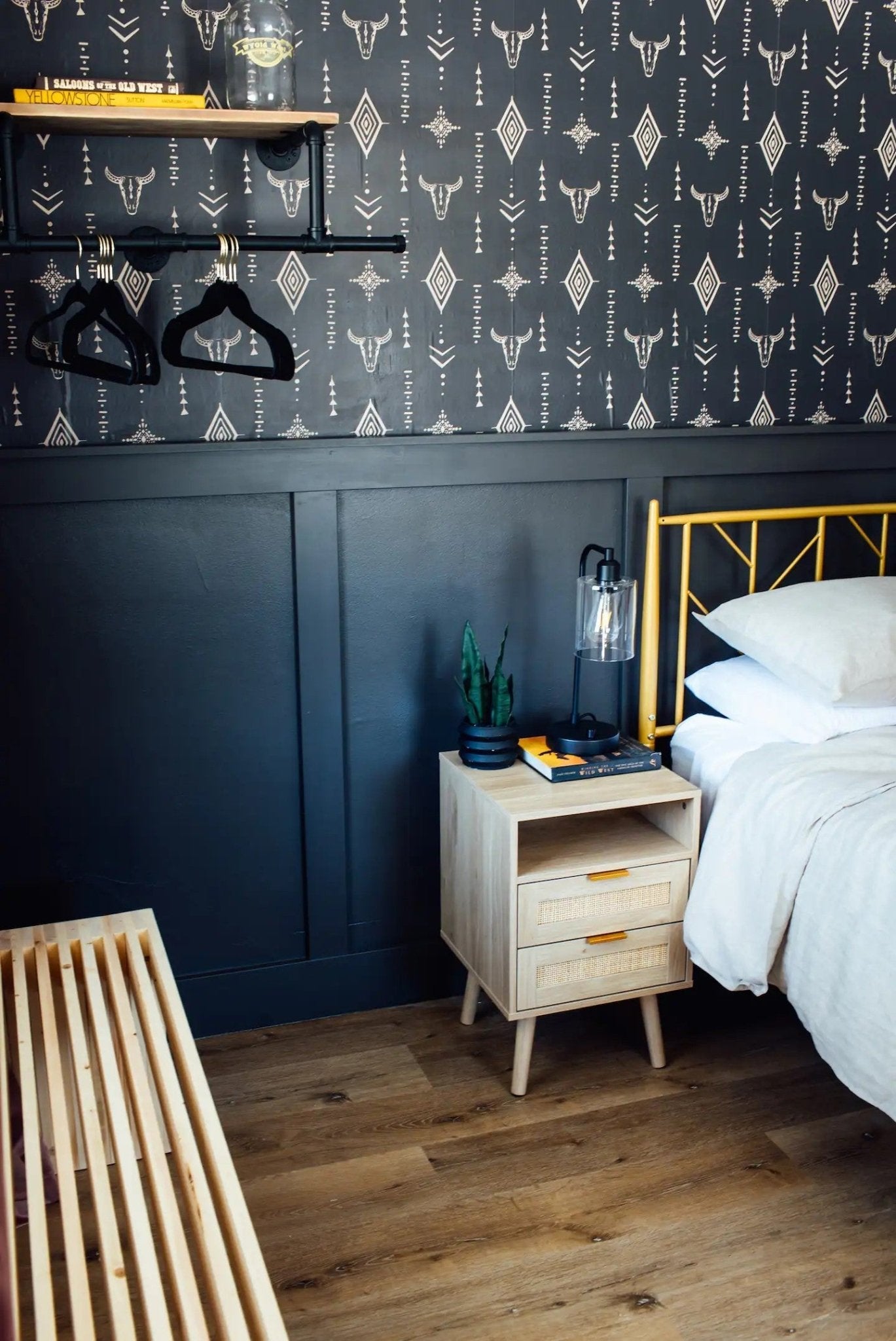  Another angle of the bedroom highlighting the Western-inspired wallpaper complemented by a dark blue paneled wall below. Beside the bed with crisp white linens is a natural wood bedside table with rattan drawer fronts, topped with a modern lantern-style lamp, books, and a small potted plant.