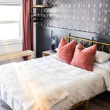 A stylish bedroom showcasing a bold Western-themed wallpaper with white illustrations of arrows, diamonds, and bull skulls on a dark background. The room features a gold metal bed frame, white bedding with coral throw pillows, and a beige throw at the foot of the bed. A dark paneled wall anchors a floating wooden shelf and a black modern chandelier adds a touch of elegance